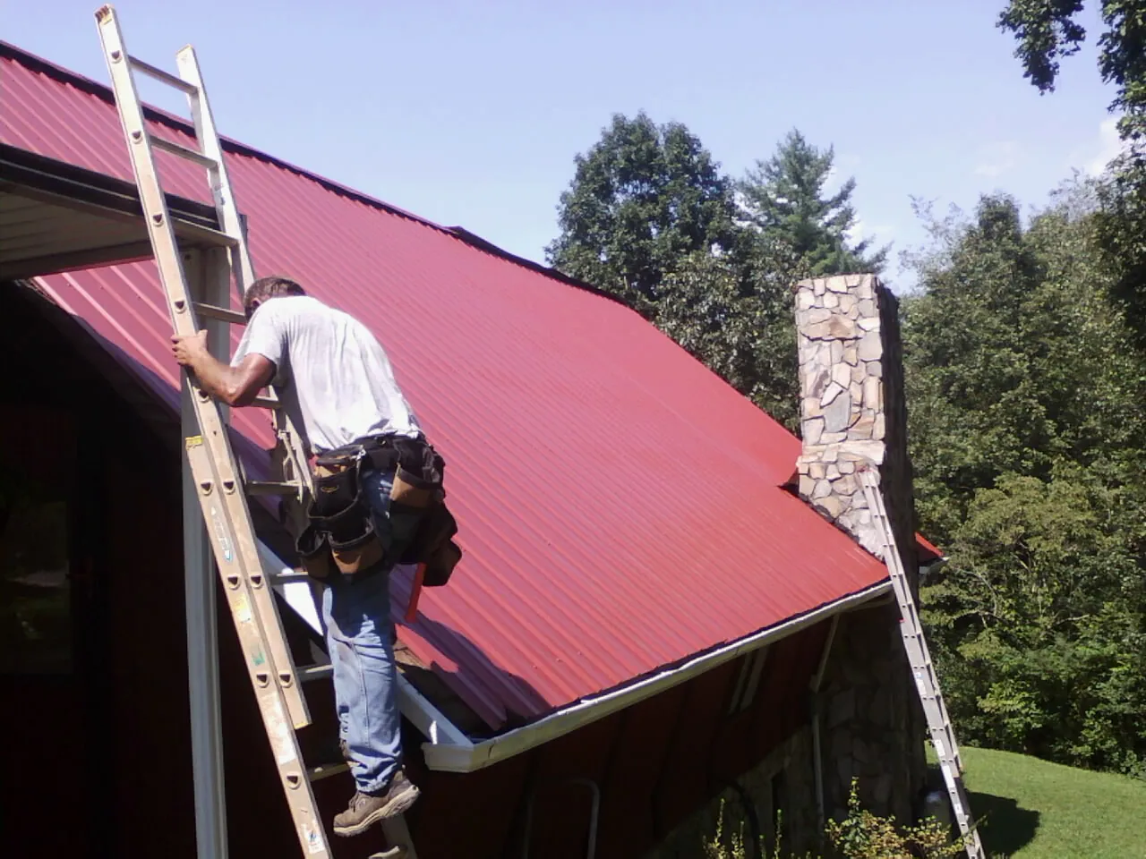 Roofer on a ladder beside a red metal roof and stone chimney — Soffit & Fascia Repair by Roofing Brothers in Cabot